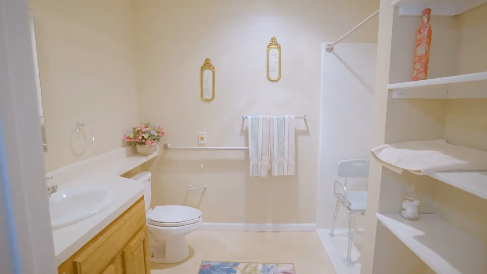 A clean and well-lit bathroom with a white toilet, a sink with a wooden cabinet, a towel rack with two towels, a shower area with a white shower chair, and shelves holding a vase and some towels. There are two decorative framed pieces on the wall and a small floral arrangement on the counter.