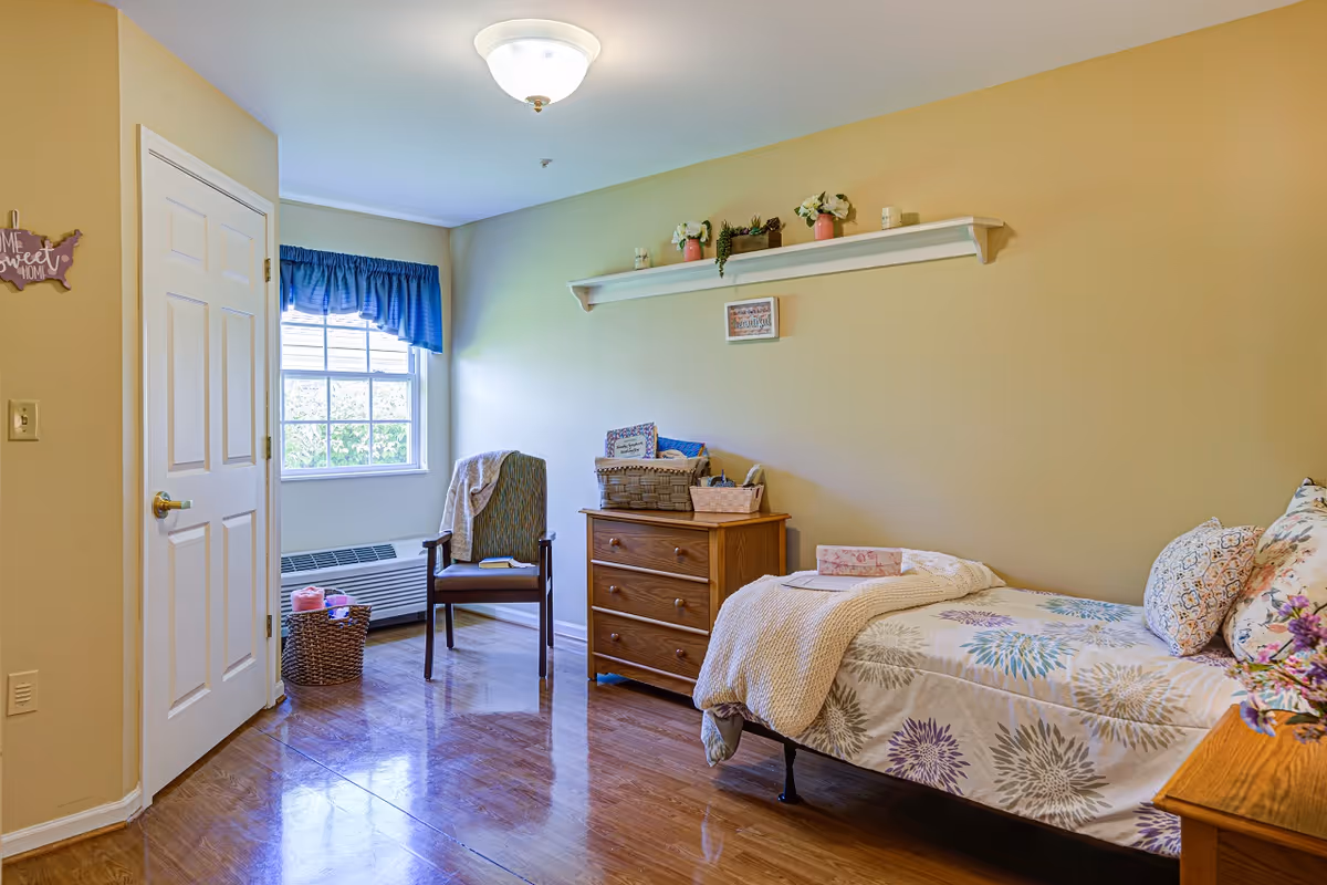 A cozy bedroom in a senior living facility with a single bed covered in floral patterned bedding and pillows. Next to the bed is a wooden nightstand with a vase of flowers. Across from the bed is a wooden dresser with baskets and decorative items on top. A wooden chair with a blanket draped over it is positioned near a window with blue curtains. The walls are painted light yellow and there is a shelf with small plants and candles above the dresser. The floor is wooden and there is a closed white door on the left side of the room.