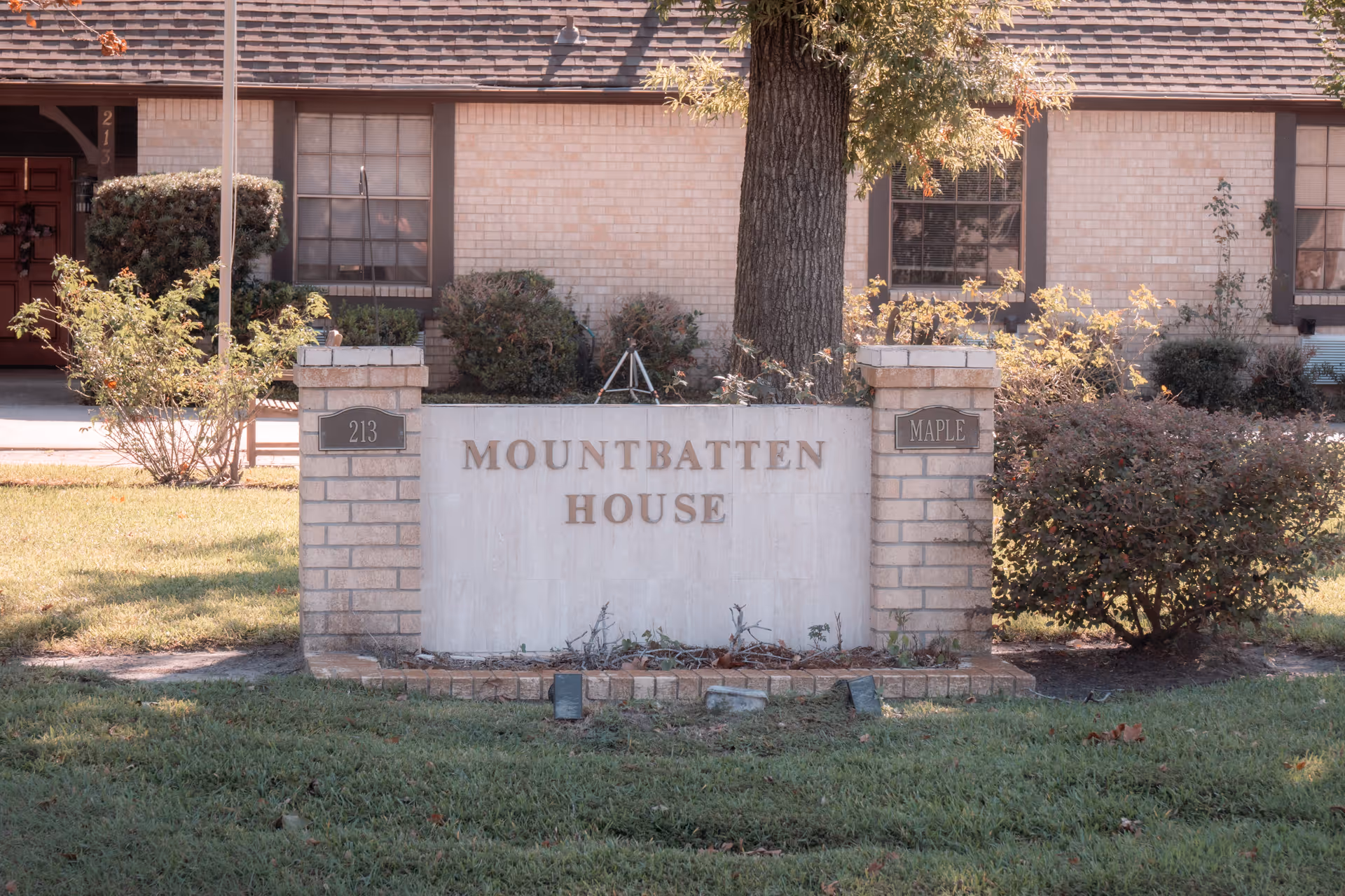 A brick sign in front of a building with the text 'Mountbatten House'. The sign is flanked by two brick pillars with plaques reading '213' and 'Maple'. There are bushes and a tree behind the sign, and the building has beige brick walls and windows with dark frames.