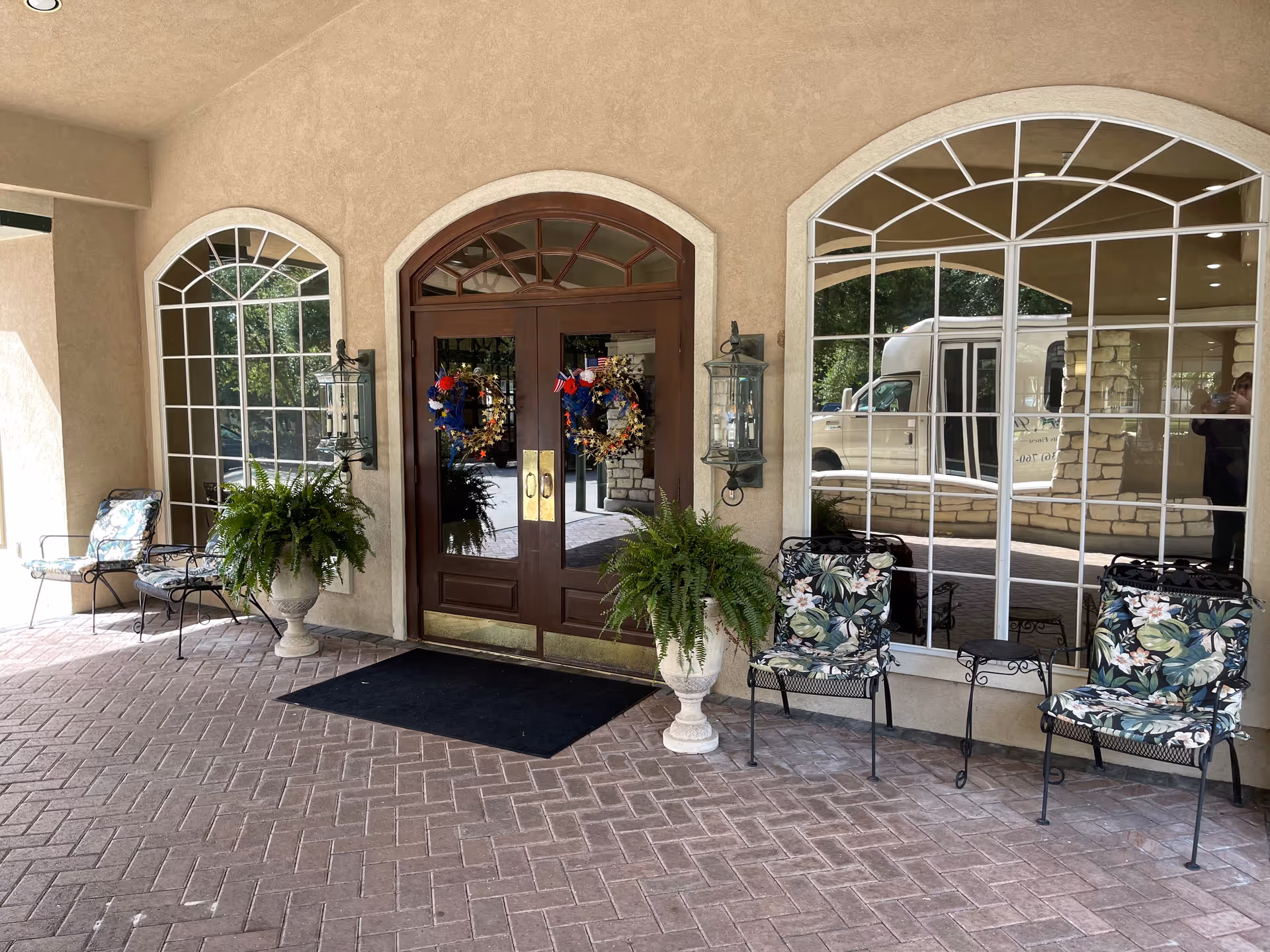 Entrance area of a facility with double wooden doors decorated with wreaths. There are large arched windows on either side of the doors, two potted ferns, and several metal chairs with floral cushions arranged around small tables on a brick-paved floor.