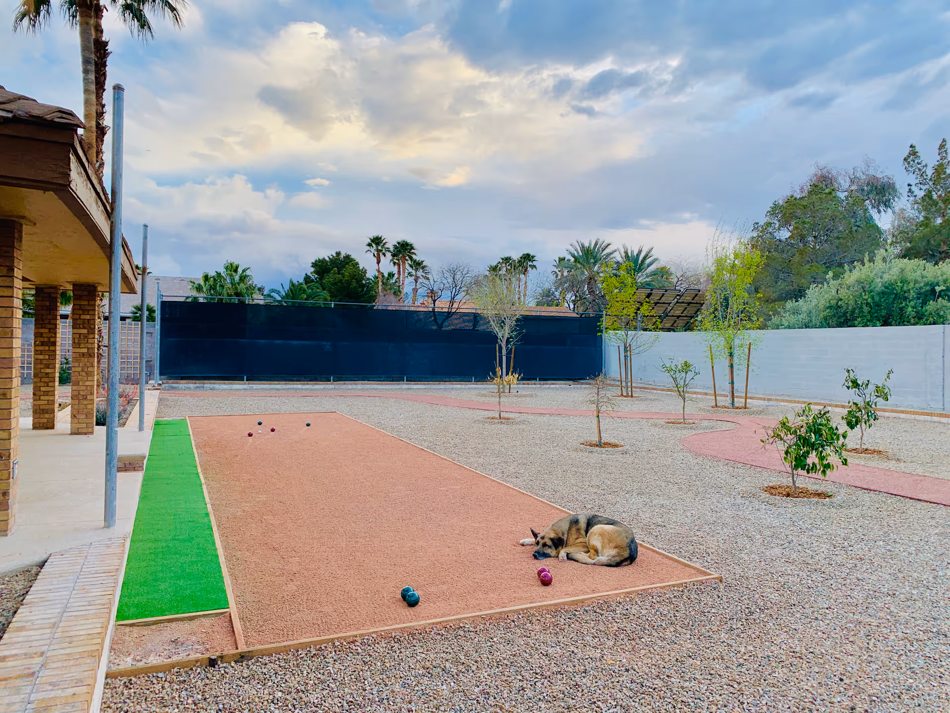 Gravel courtyard with a bocce-style court, small newly planted trees, scattered toys, and a dog lying on the court beside a covered patio.