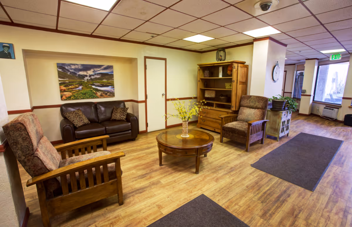 A cozy seating area in a senior living facility with two upholstered armchairs and a dark brown leather loveseat arranged around a round wooden coffee table with a vase of yellow flowers. The room has wooden flooring, a wooden cabinet with shelves, a wall clock, and a large window letting in natural light.
