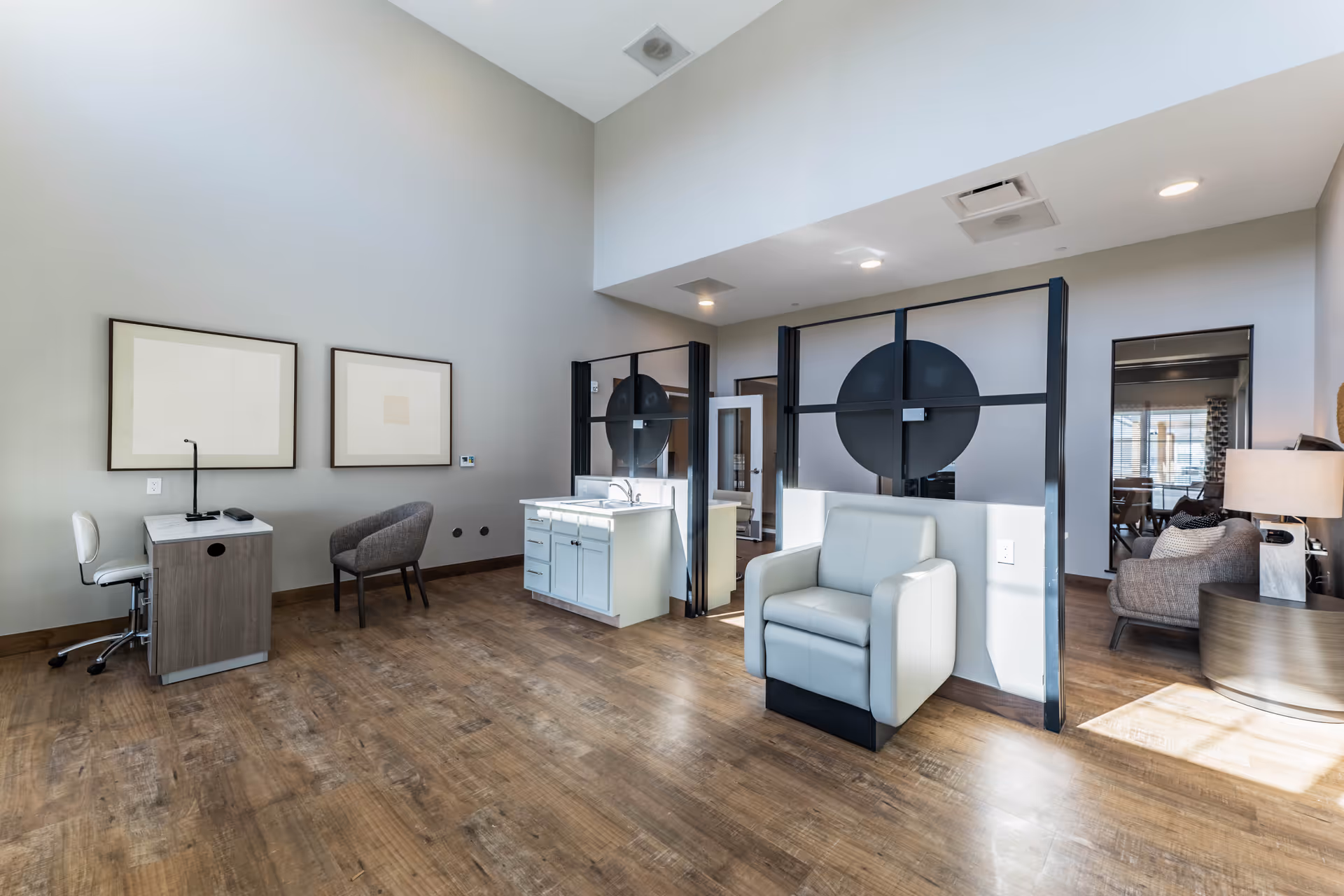 A modern, spacious interior room with high ceilings and wood flooring. The room features a small desk with a white chair and a gray armchair against a light gray wall with two framed abstract artworks. There is a white cabinet with a sink in the center, partially dividing the space with black-framed glass partitions. On the right side, there is a light gray armchair and a glimpse of a cozy seating area with a sofa, a lamp, and a round side table near a window with blinds.