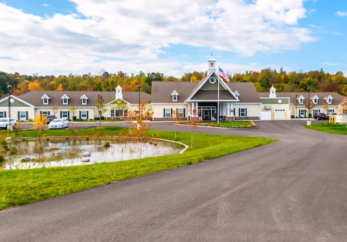 Exterior view of a single-story senior living facility building with a large driveway, a small pond with some rocks and young trees in front, and an American flag on a flagpole near the entrance. The building has a beige and green color scheme with multiple windows and dormers on the roof. Trees with autumn foliage are visible in the background under a partly cloudy sky.