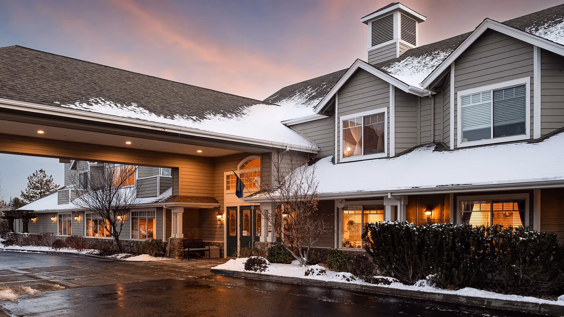 Exterior view of Atria Summit Ridge senior living facility at dusk with snow on the roof and ground, warm lights glowing from windows, and a covered entrance area.