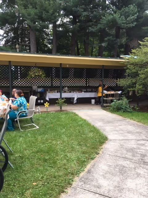 Outdoor covered patio area with tables set up for dining. Several people are seated at a table on the left side, and a person in a yellow shirt is standing near a buffet table under the covered area. The area is surrounded by green grass, trees, and bushes.