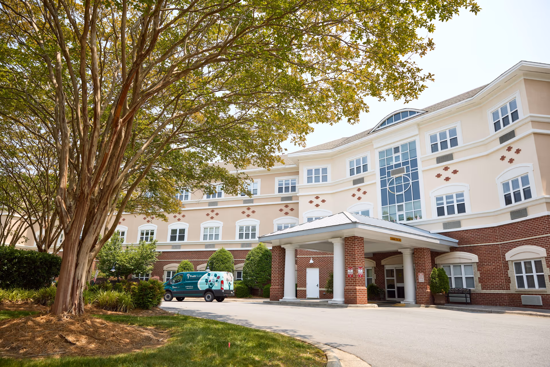 Exterior view of a multi-story senior living facility building with a covered entrance supported by white columns. There is a large tree with green leaves on the left side and a green van parked near the entrance. The building has a combination of brick and light-colored walls with multiple windows.