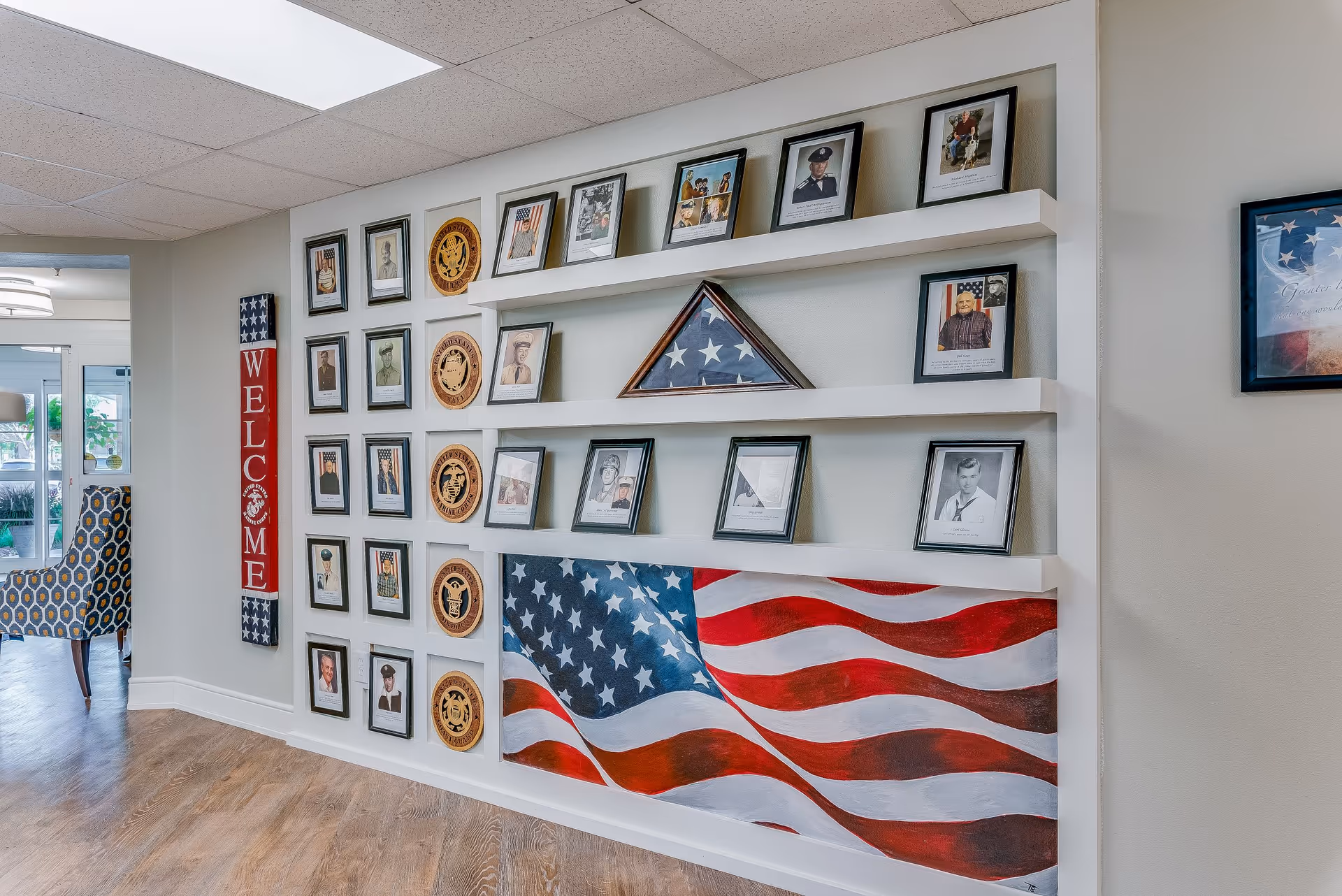Interior wall display in a senior living facility featuring framed photographs of veterans, military insignias, a folded American flag in a triangular case, and a large painted American flag. A vertical wooden sign with the word 'WELCOME' is mounted on the left side of the wall. Part of a patterned chair and a doorway leading to another room are visible on the left.