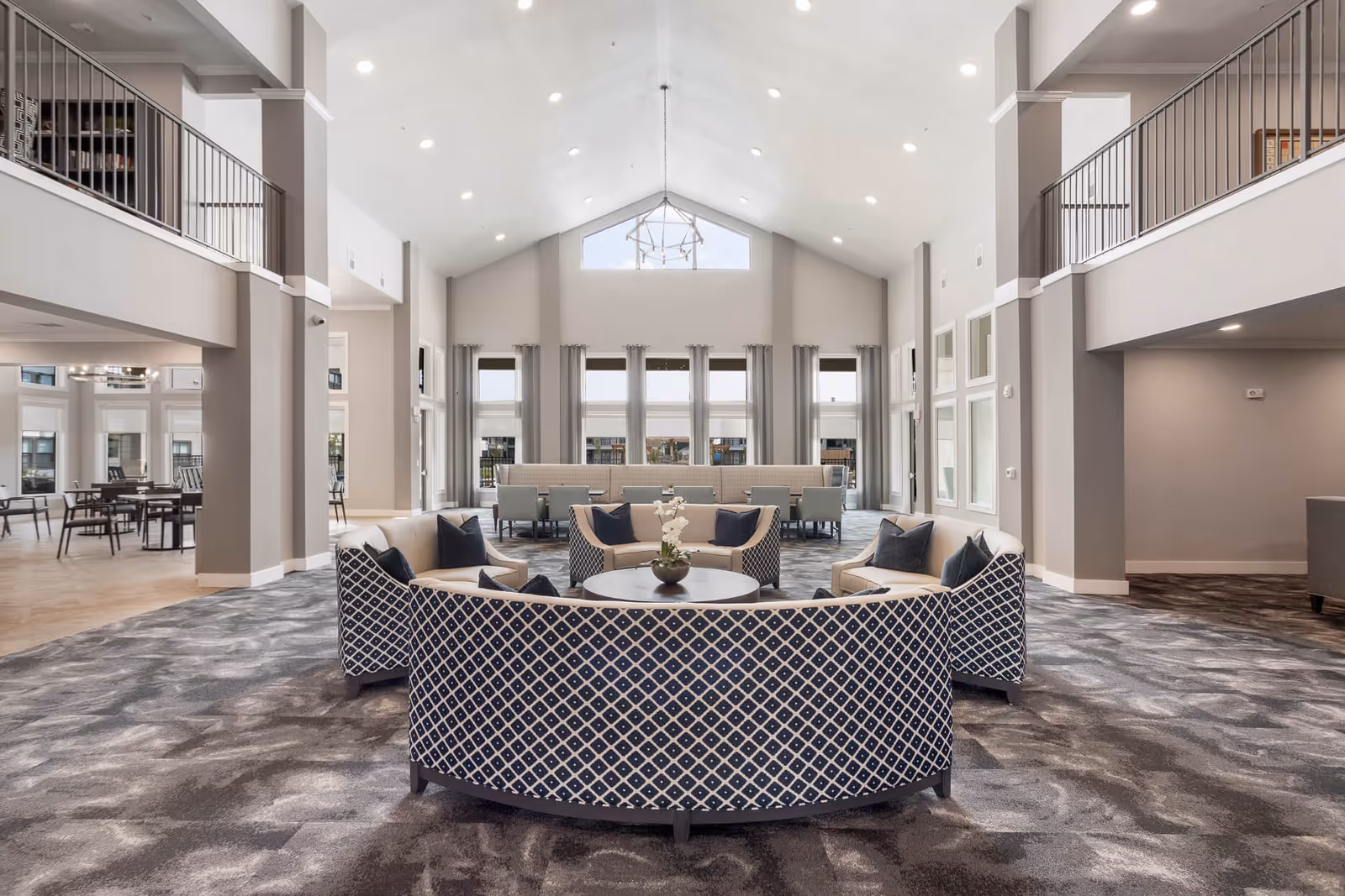Spacious two-story communal lounge with curved patterned sofas around a central coffee table, high vaulted ceiling, large windows, and upper balconies.