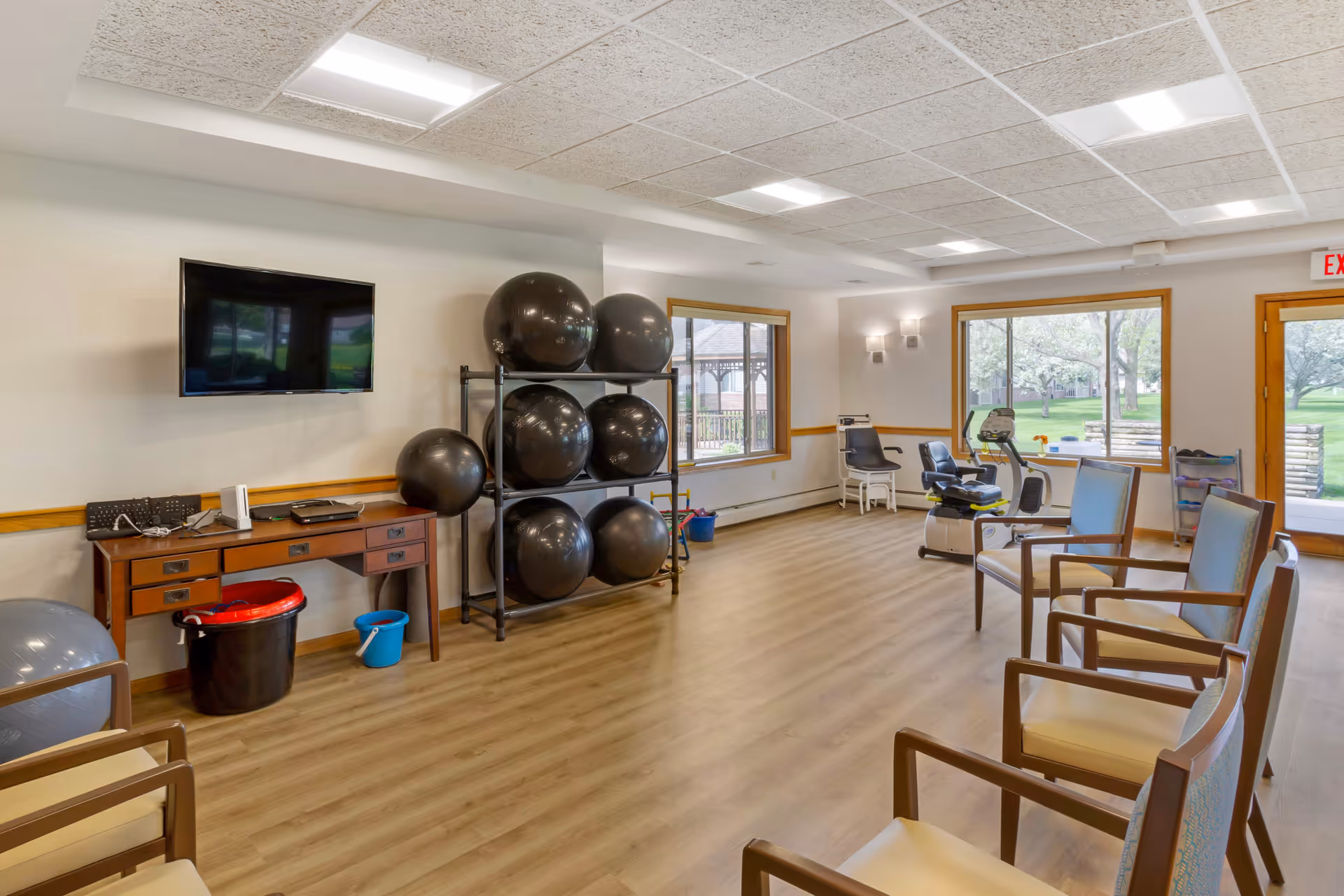 A spacious senior living exercise room with wooden flooring, several large black exercise balls stacked on a metal rack, a stationary exercise bike, chairs arranged along the walls, a wall-mounted flat screen TV, and large windows showing a green outdoor area.