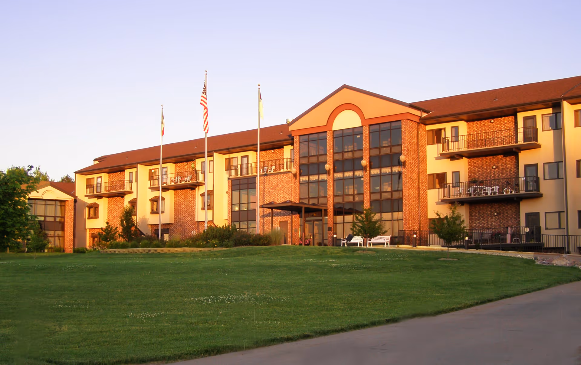 Exterior view of a multi-story senior living facility building with brick and beige walls, several balconies, large windows, and three flagpoles with flags in front. The building is surrounded by a well-maintained green lawn and a paved walkway.