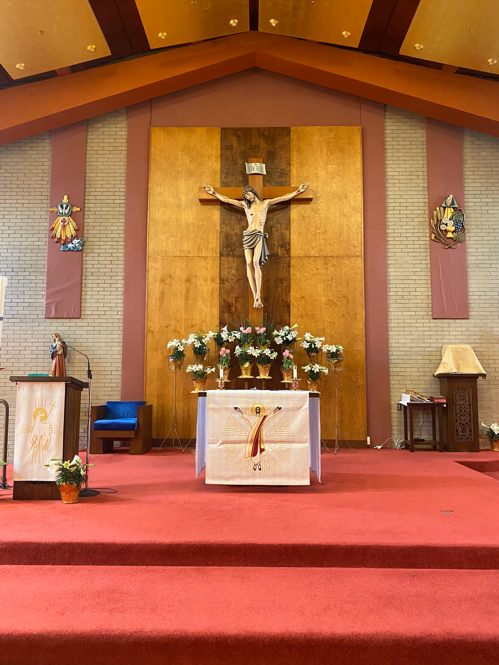 Interior view of a church sanctuary showing a crucifix mounted on a wooden panel above an altar draped with a white cloth and surrounded by floral arrangements on red carpeted steps.