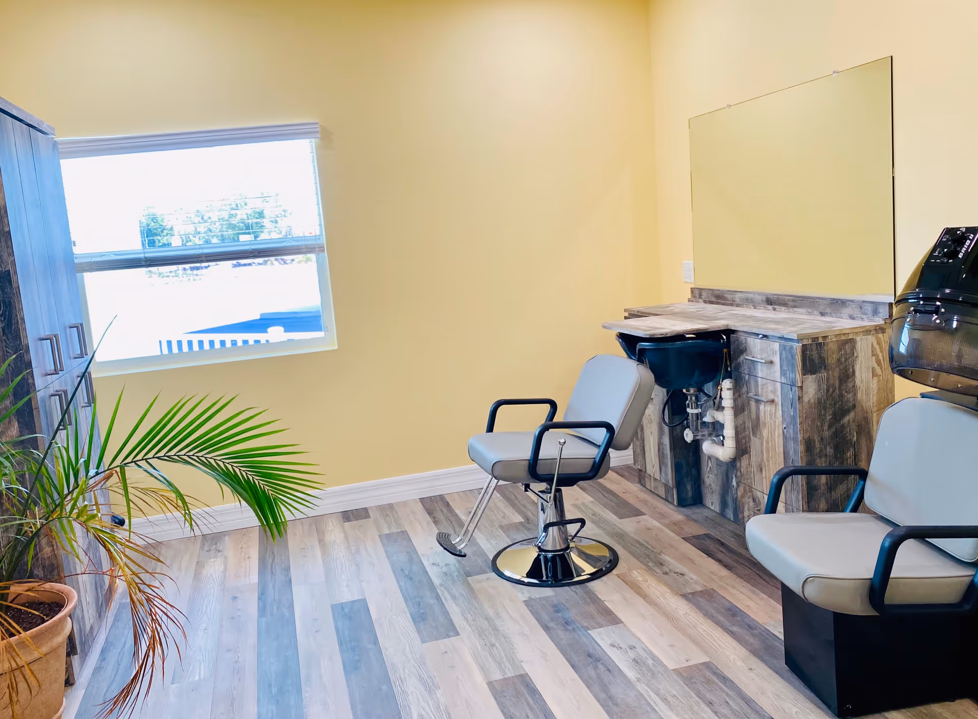 Bright salon-style room with two styling chairs, a shampoo sink and mirror, wood-look floors, and a potted plant by a window.