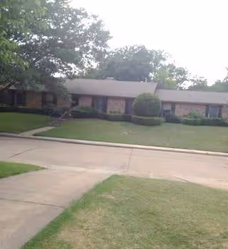 Single-story residential building with a brick exterior, surrounded by trimmed bushes and trees, viewed from across a paved street with a grassy lawn in the foreground.