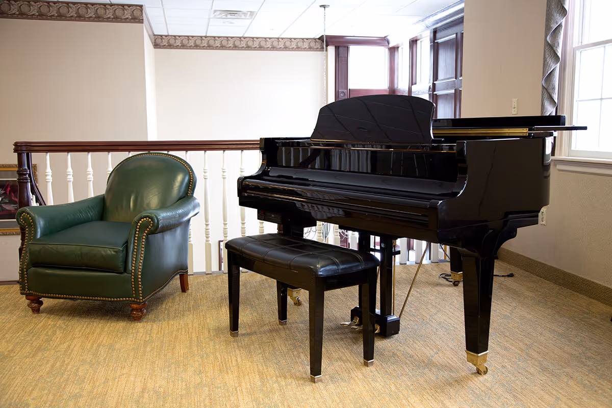 A black grand piano with a matching black bench is placed on a carpeted floor near a window. To the left of the piano, there is a green leather armchair with wooden legs and brass nailhead trim. The room has beige walls with a decorative border near the ceiling and white railing in the background.
