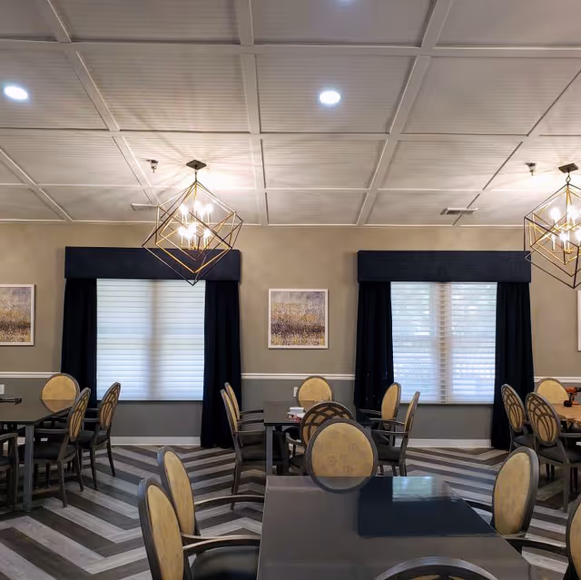 Interior view of a dining room with multiple tables and chairs arranged neatly. The room features large windows with white blinds and dark curtains, modern geometric pendant lights hanging from a white paneled ceiling, and framed artwork on beige walls. The floor has a chevron pattern in shades of gray and white.