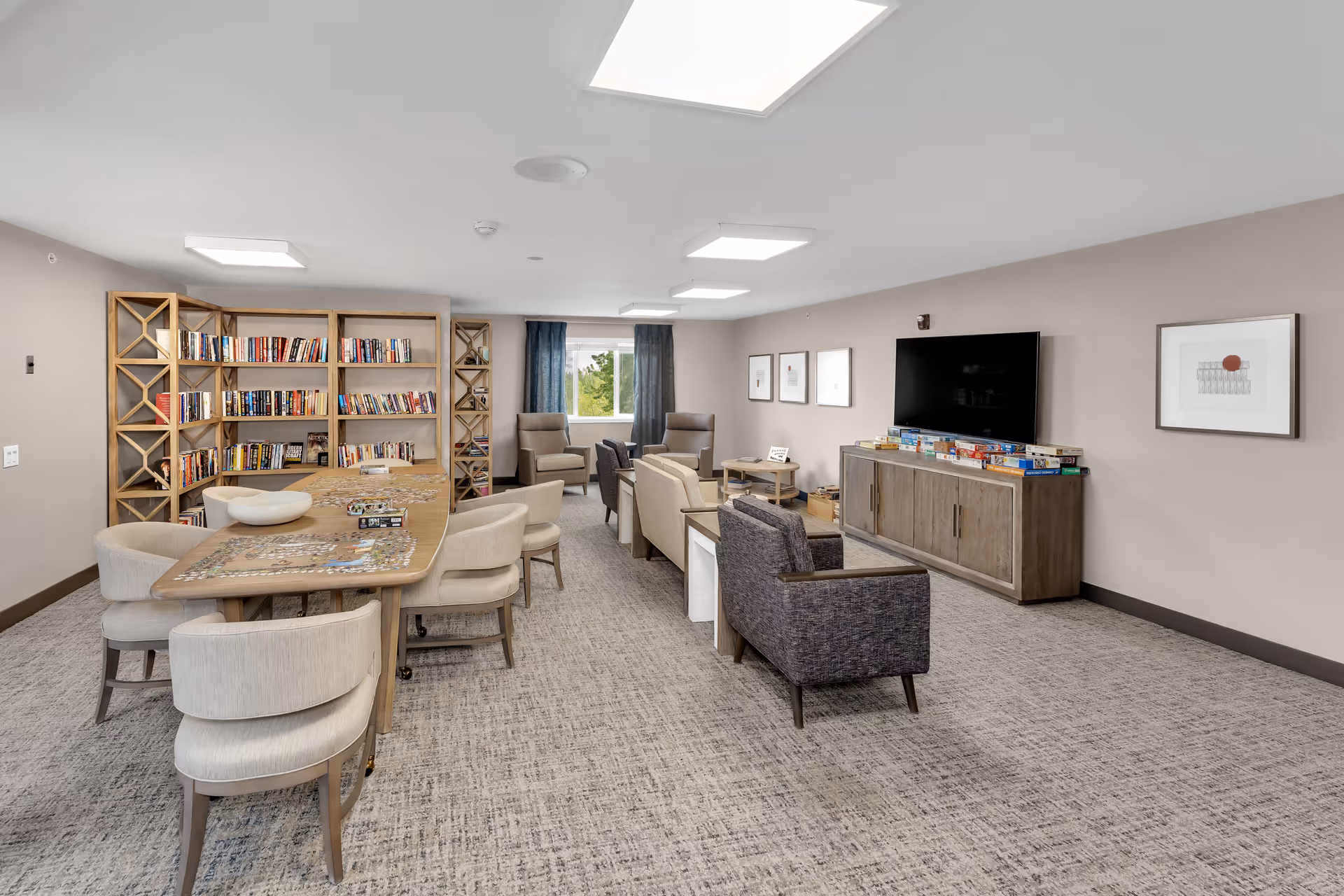 Bright community lounge with a large table and chairs, bookshelves, a seating area, and a wall-mounted TV.