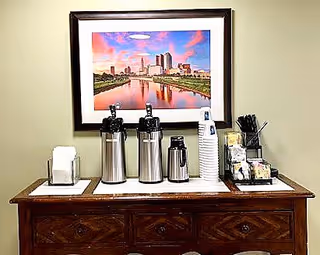 A wooden sideboard with three stainless steel coffee dispensers, a stack of white disposable cups, napkins, and a container holding sugar packets and stirrers. Above the sideboard is a framed picture of a city skyline at sunset with a river reflecting the buildings.