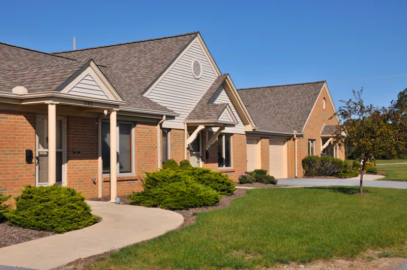 Exterior view of a single-story senior living community building with brick walls, beige siding, and multiple entrances. The building is surrounded by green grass, small bushes, and a clear blue sky.