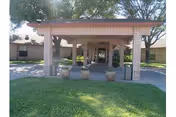 Entrance area of La Vida Serena Nursing & Rehabilitation facility with a covered walkway supported by brick columns, three large potted plants, and a well-maintained lawn in the foreground. Trees and parts of the building are visible in the background.