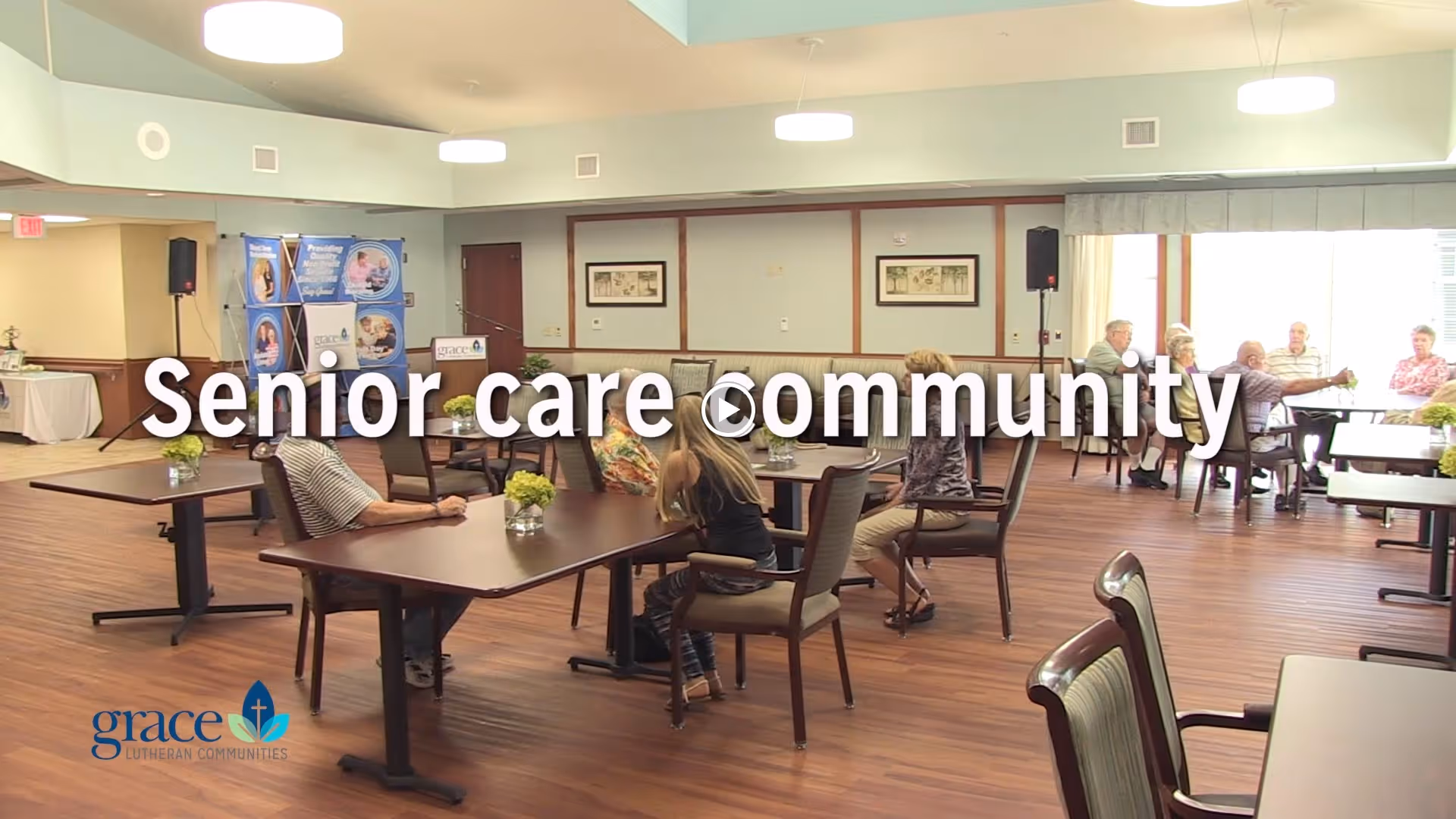 A spacious dining area in a senior care community with several elderly residents seated at tables. The room has wooden flooring, light-colored walls, and large windows allowing natural light to enter. Tables are decorated with small flower arrangements. There are informational banners and a podium in the background.