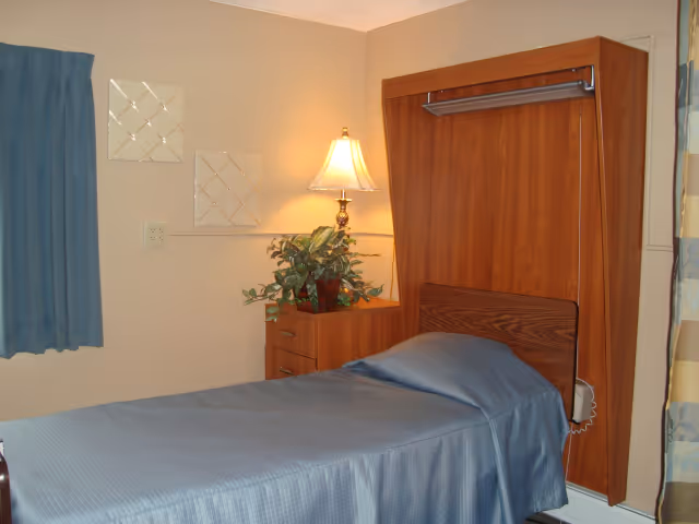 A single bed with a blue bedspread in a room with beige walls. Next to the bed is a wooden nightstand with a potted plant and a lamp. There are two decorative wall hangings above the nightstand and a blue curtain on the window to the left.