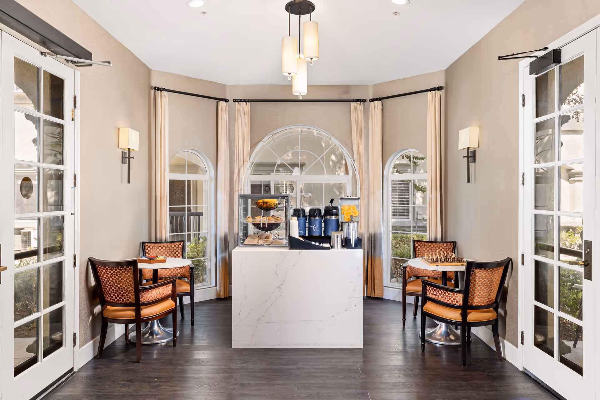 Dining nook with two small round tables and upholstered chairs flanking a central marble beverage station beneath arched windows.