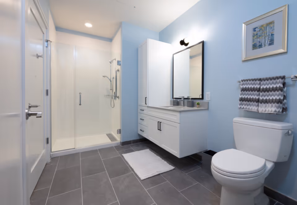 Modern bathroom with a glass walk-in shower, floating vanity and mirror, and a toilet against light blue walls.