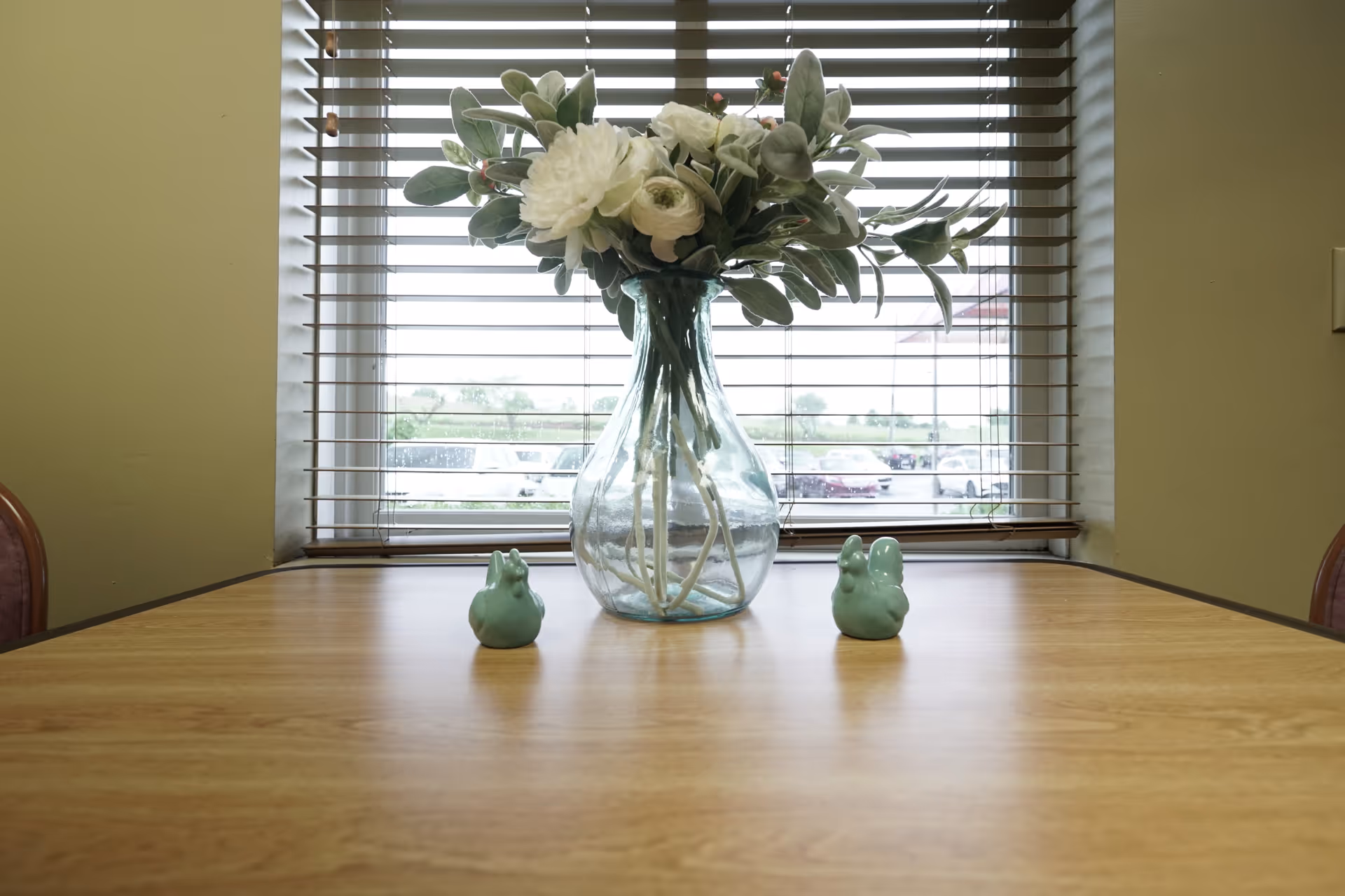 A wooden table with a clear glass vase holding white flowers and green leaves placed in front of a window with horizontal blinds. Two small green ceramic bird figurines are positioned on either side of the vase. The background shows a parking lot and some greenery outside the window.