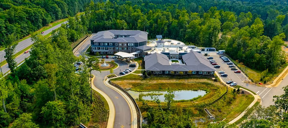 Aerial view of a senior living facility named Watercrest Richmond surrounded by dense green trees. The facility consists of multiple connected buildings with parking lots and a circular driveway featuring a fountain in the center. There is a small pond near the buildings and a road leading to the entrance.