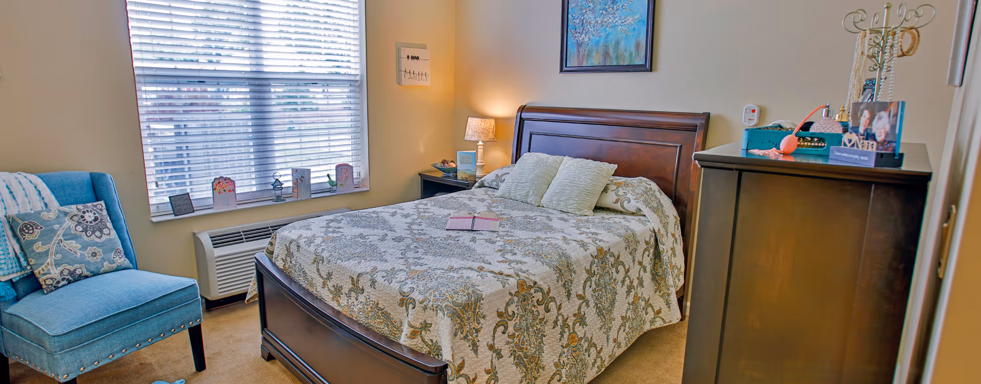 A cozy bedroom with a wooden bed frame and a patterned bedspread, two pillows, a blue upholstered chair with a decorative pillow and blanket, a window with blinds, a nightstand with a lamp, and a dresser with personal items and photos on top. A colorful painting hangs on the wall above the bed.