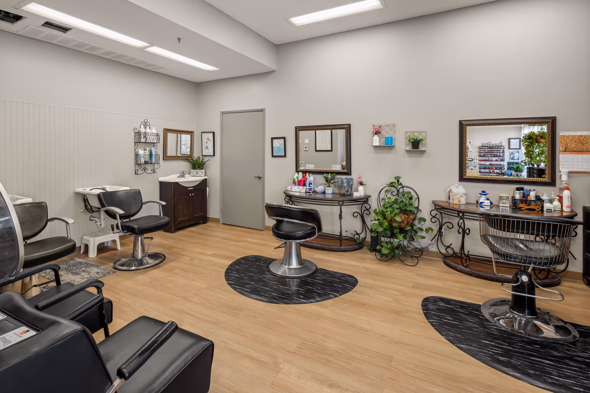 Interior view of a senior living community hair salon with multiple styling chairs, mirrors, a hair washing station, and shelves with hair care products. The room has light-colored walls, wood flooring, and decorative plants.