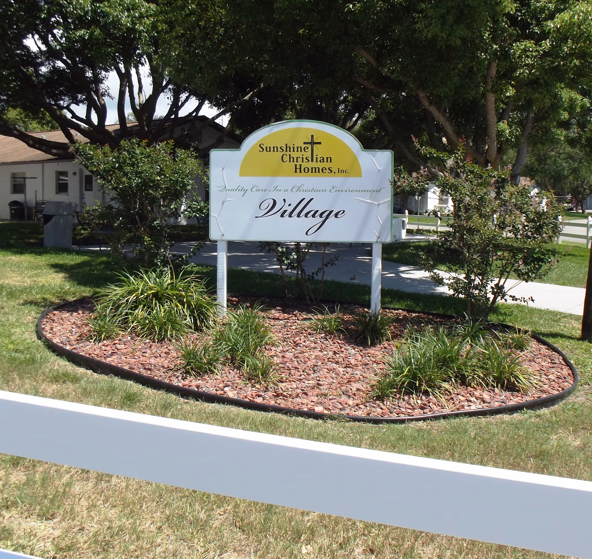 Outdoor view of a landscaped area with a white sign that reads 'Sunshine Christian Homes, Inc. Quality Care In a Christian Environment Village' surrounded by trees, bushes, and grass with a white fence in the foreground.