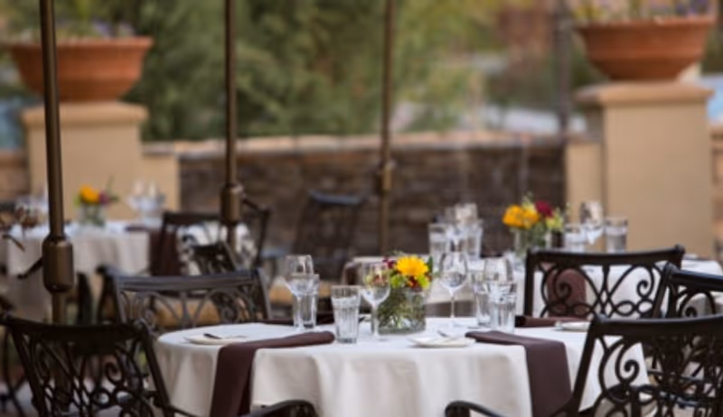 Outdoor dining area with round tables covered in white tablecloths and brown napkins, set with glasses, plates, and small flower arrangements. Wrought iron chairs surround the tables, and large planters and greenery are visible in the background.