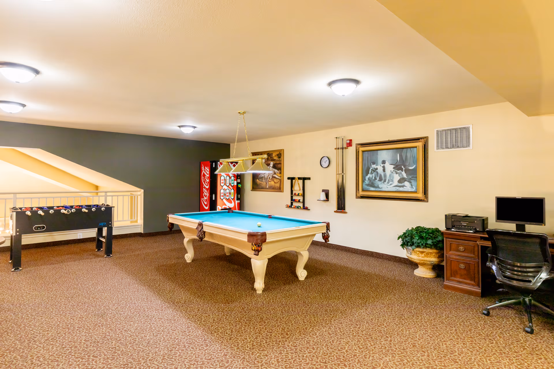 A recreational room featuring a pool table with a single cue ball, a foosball table, a Coca-Cola vending machine, a wall-mounted rack with pool cues and balls, a framed painting of dogs, a clock, a potted plant, and a desk with a computer and printer.
