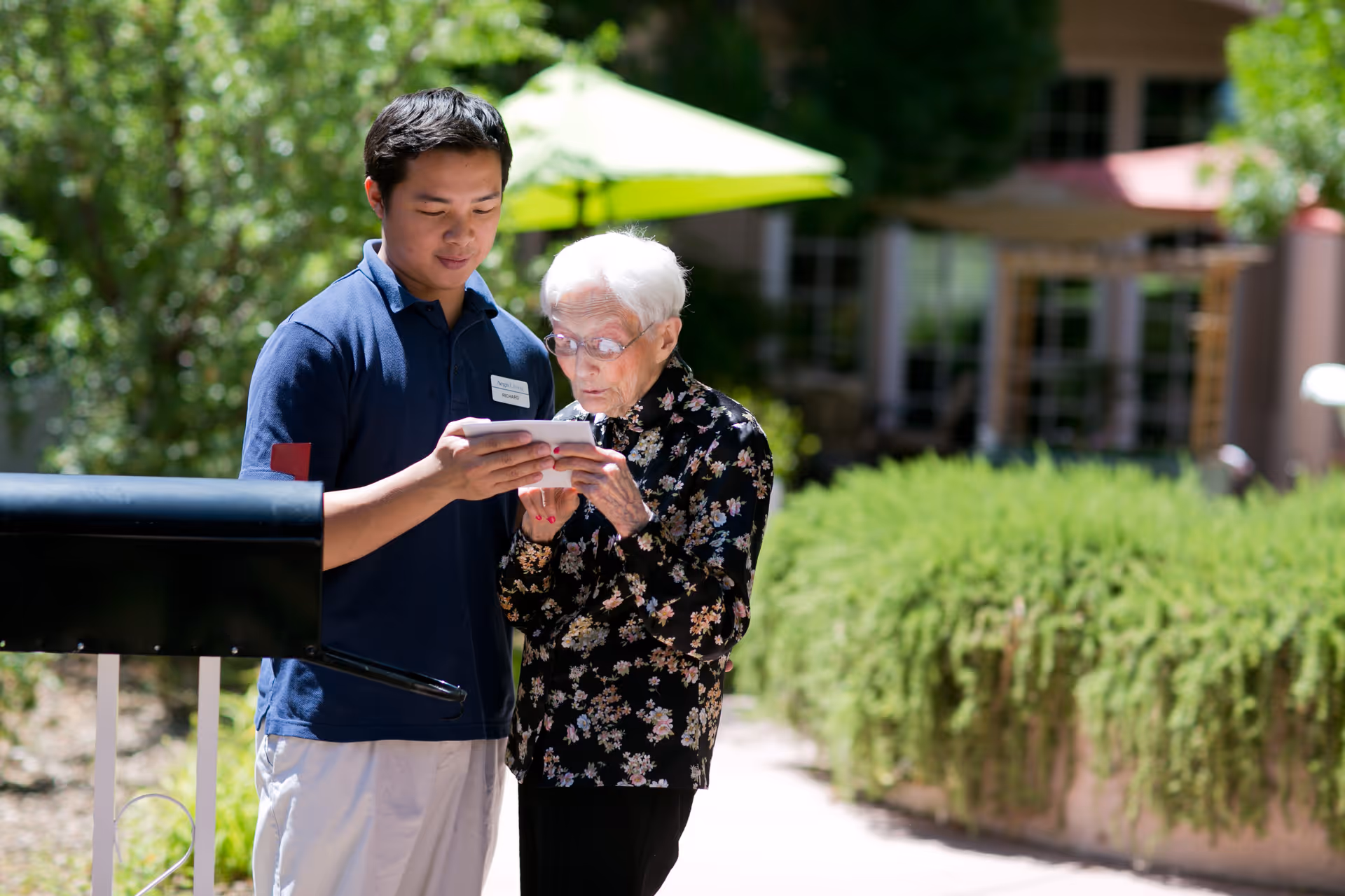 A young man in a navy blue shirt is assisting an elderly woman with white hair and glasses as they both look at a smartphone outdoors near a mailbox, with greenery and a building in the background.