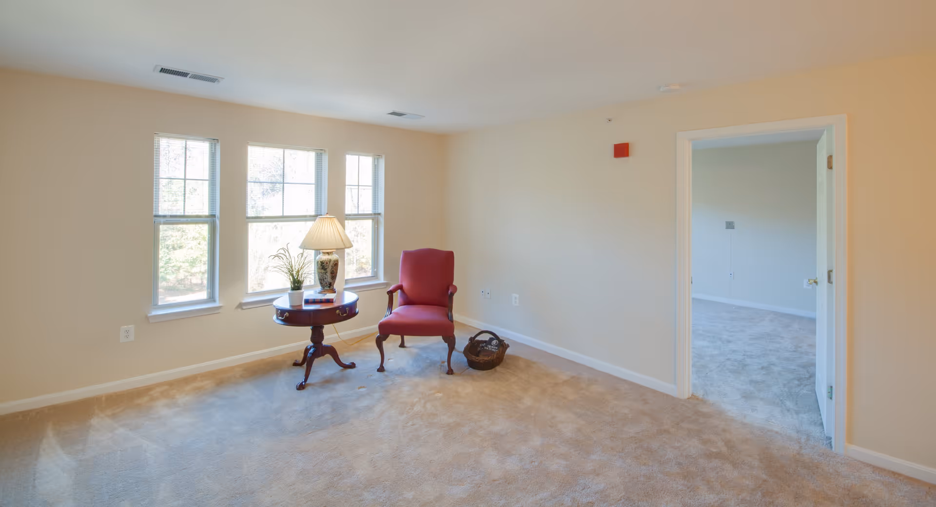 A bright room with beige walls and carpeted floor featuring three large windows letting in natural light. There is a small round wooden table with a lamp, a plant, and a book on it, next to a red upholstered armchair. A basket is placed on the floor near the chair. An open doorway leads to another carpeted room.