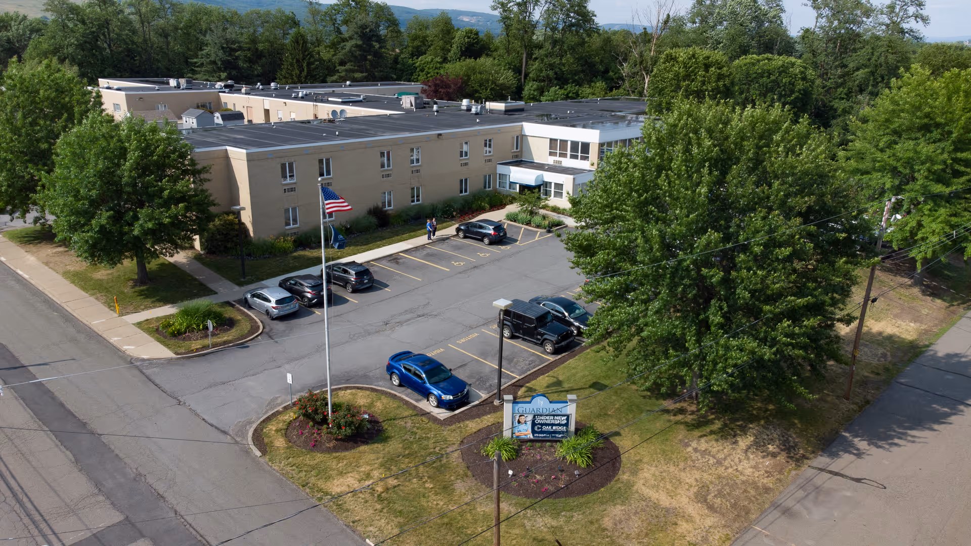 Aerial view of a two-story beige rehabilitation and healthcare building with a parking lot, flagpole, landscaping, and an entrance sign.