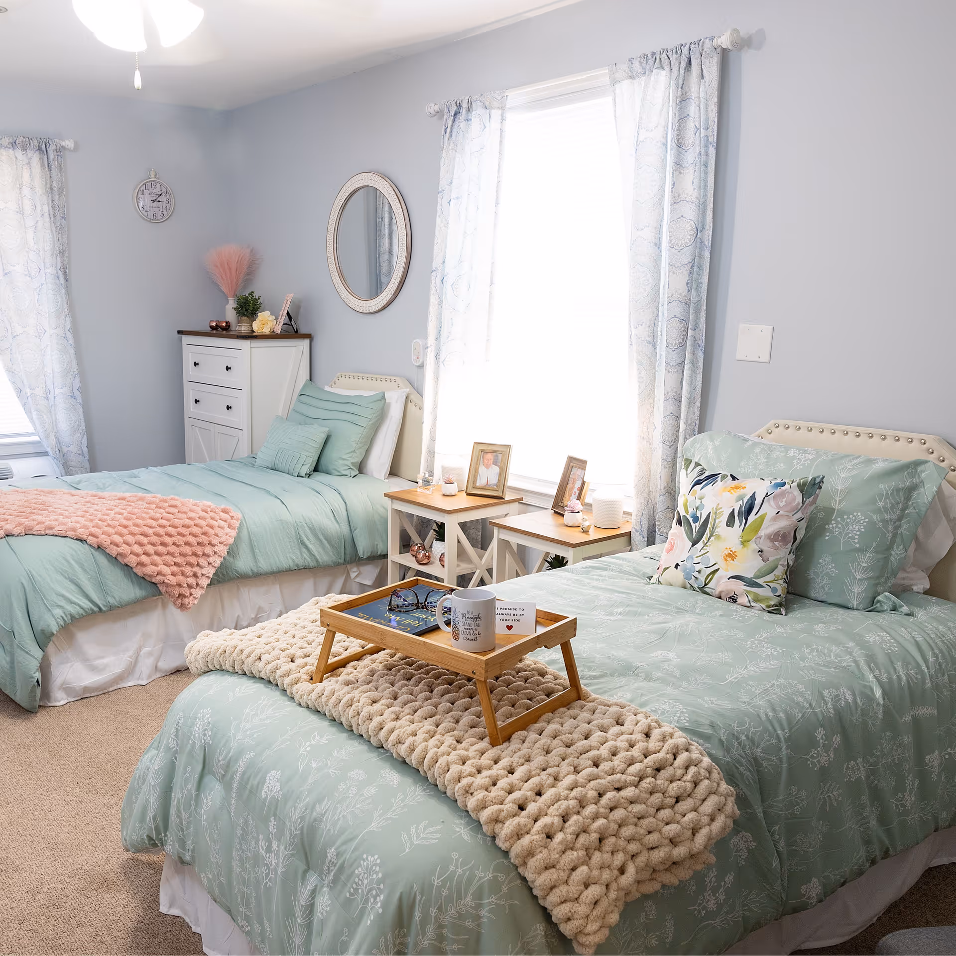 A cozy bedroom with two twin beds dressed in light green bedding with floral patterns. One bed has a chunky beige knit blanket and a wooden tray holding a mug, glasses, and a small card. The other bed has a pink textured throw blanket. Between the beds are two small white nightstands with framed photos, candles, and small decorative items. The walls are painted light gray with two windows covered by light patterned curtains. A white dresser with drawers and decorative items is against the wall, along with a round mirror and a wall clock.