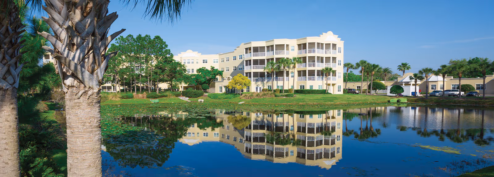 A large, multi-story beige senior living facility building named Cypress Palms is reflected in a calm pond surrounded by green grass, palm trees, and other vegetation under a clear blue sky.