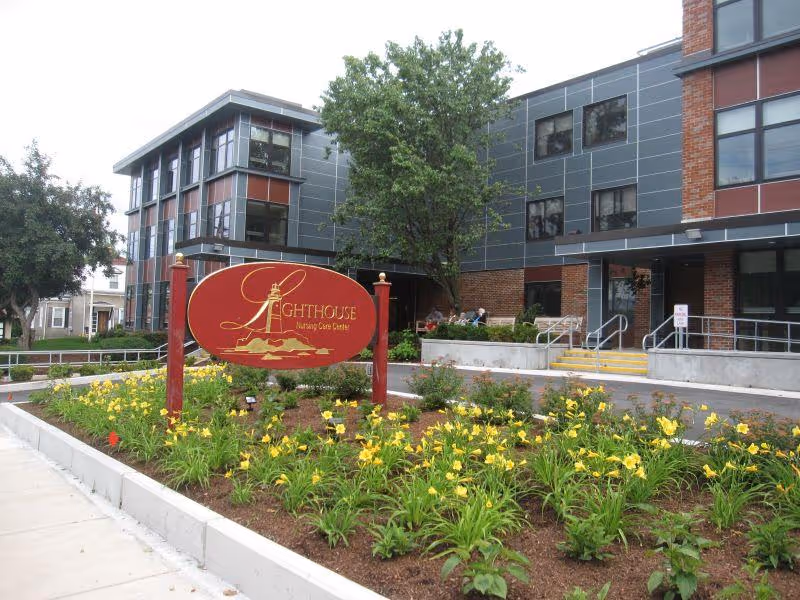 Exterior view of Lighthouse Rehabilitation & Healthcare Center showing a modern three-story building with large windows, a landscaped garden with yellow flowers, and a red sign with a lighthouse logo and the facility name.