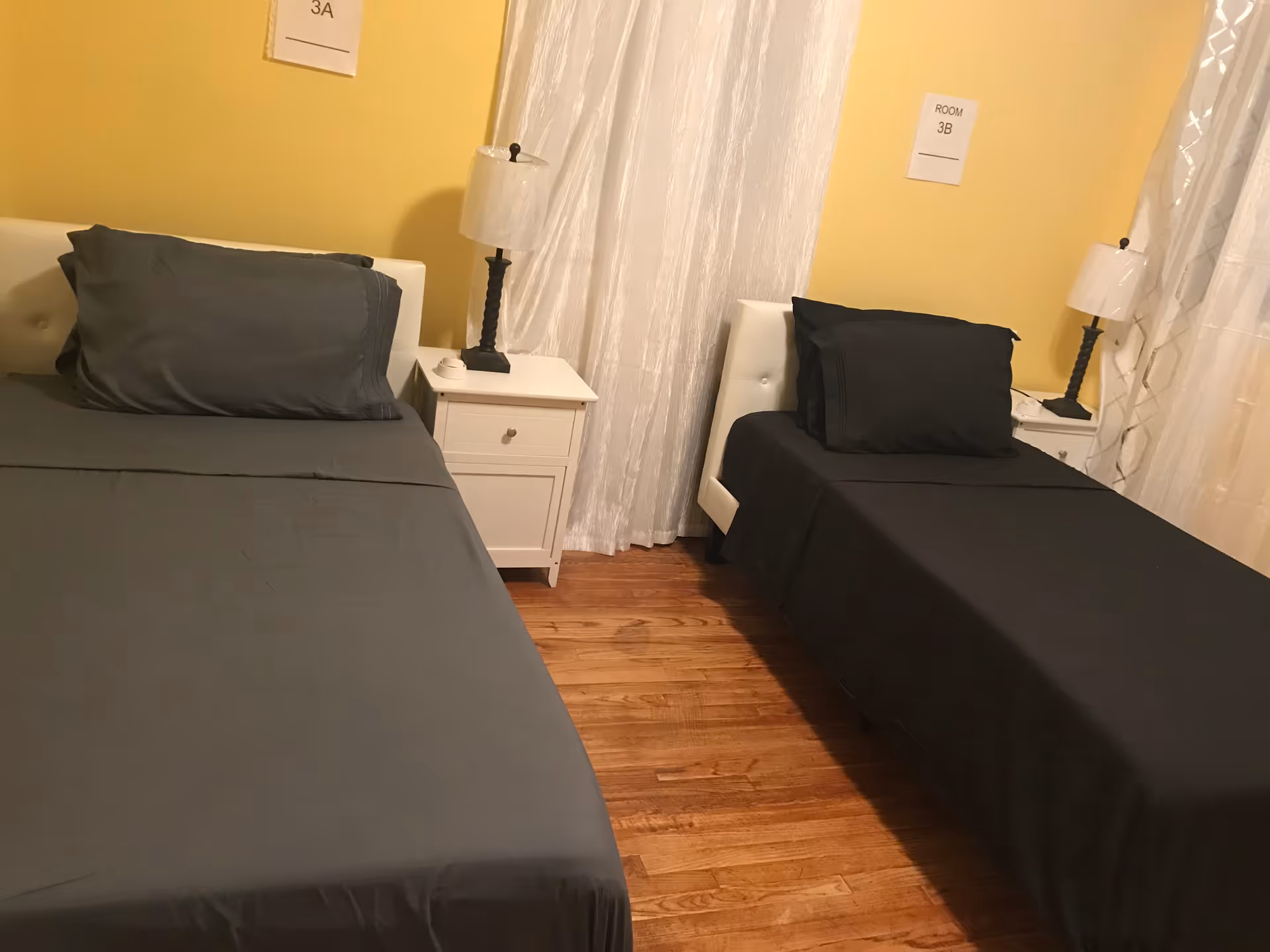 A bedroom with two beds, one larger bed on the left with gray bedding and a smaller bed on the right with black bedding. Each bed has a white headboard and a white nightstand with a lamp. The walls are painted yellow, and there are white curtains on the windows. Signs above the beds read '3A' and 'Room 3B'.