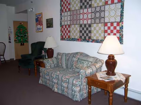 Cozy senior living lounge with a patterned sofa, two lamps, side tables, and a wall quilt.