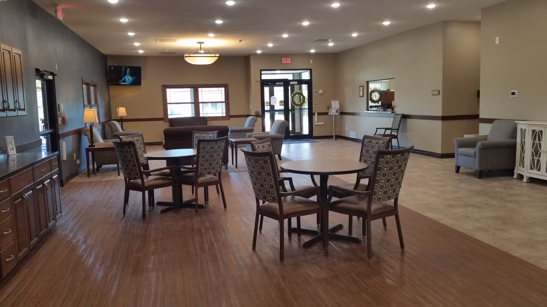 A spacious senior living facility common area with round tables and patterned chairs on a wood floor section. There are additional seating areas with armchairs and sofas near windows and doors. The walls are beige with dark wood trim, and a television is mounted on the far wall.