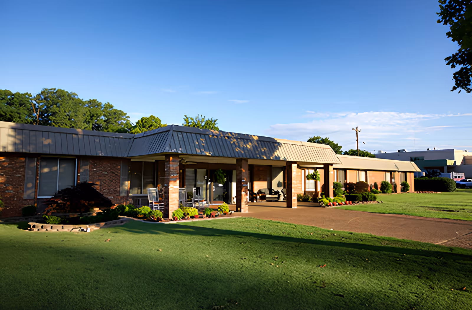 Exterior view of Ahc West Tennessee Transitional Care building with a covered entrance, brick walls, large windows, and a well-maintained lawn with shrubs and trees under a clear blue sky.