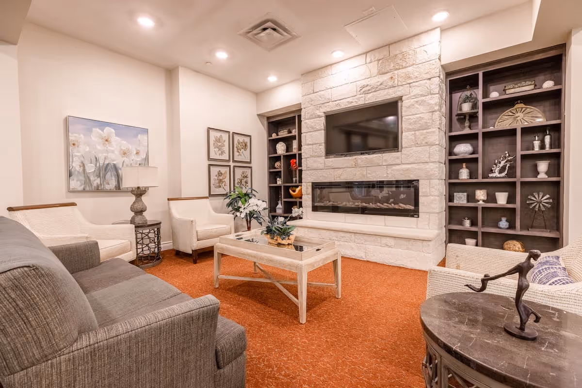 A cozy living room with a gray sofa, two white armchairs, a wooden coffee table with a plant centerpiece, and a stone fireplace with a mounted TV above it. The room has built-in dark wood shelves filled with decorative items, floral artwork on the walls, and warm lighting from recessed ceiling lights.
