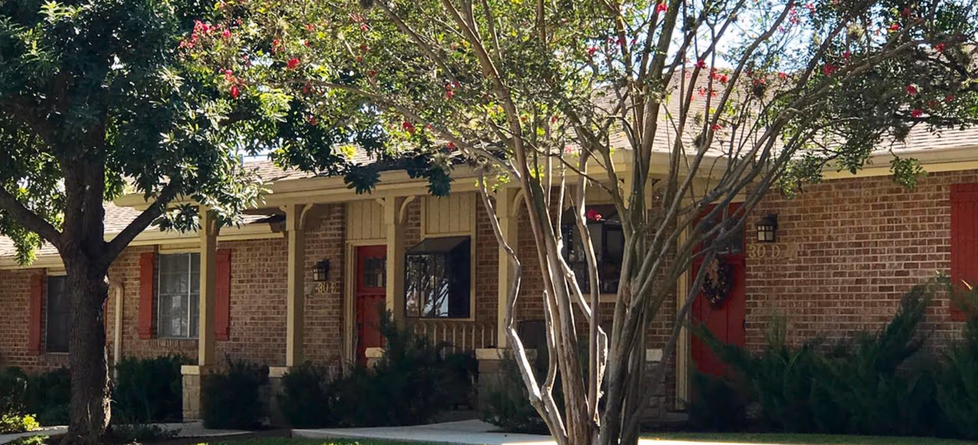 Exterior view of a single-story brick building with red doors and windows, surrounded by trees and shrubs under a clear sky.