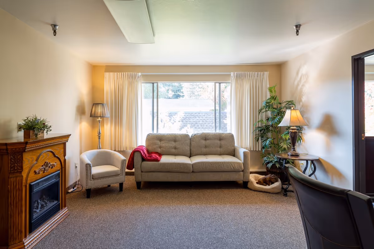 Cozy living room with a sofa, armchairs, a fireplace, lamps, and a large window letting in natural light.