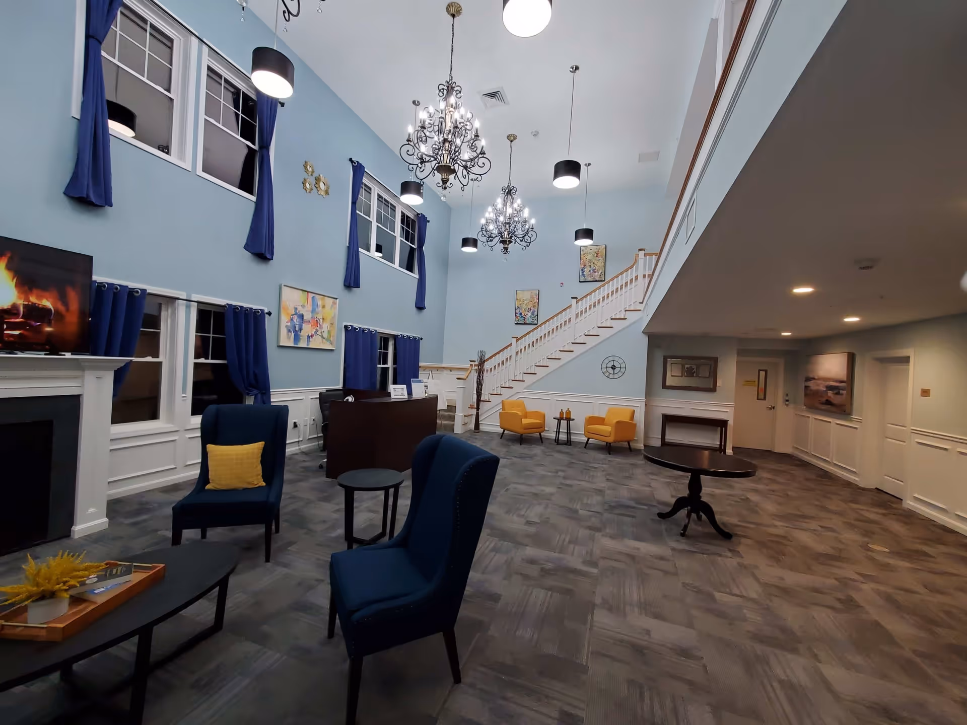 A spacious senior living facility common area with high ceilings painted light blue, featuring two ornate chandeliers and several modern pendant lights. The room has multiple windows with dark blue curtains, a fireplace with a TV above it showing a fire, and comfortable seating including blue chairs with yellow pillows and yellow armchairs near a staircase. The floor is covered with gray patterned carpet, and there are various pieces of wall art and decorative items throughout the space.