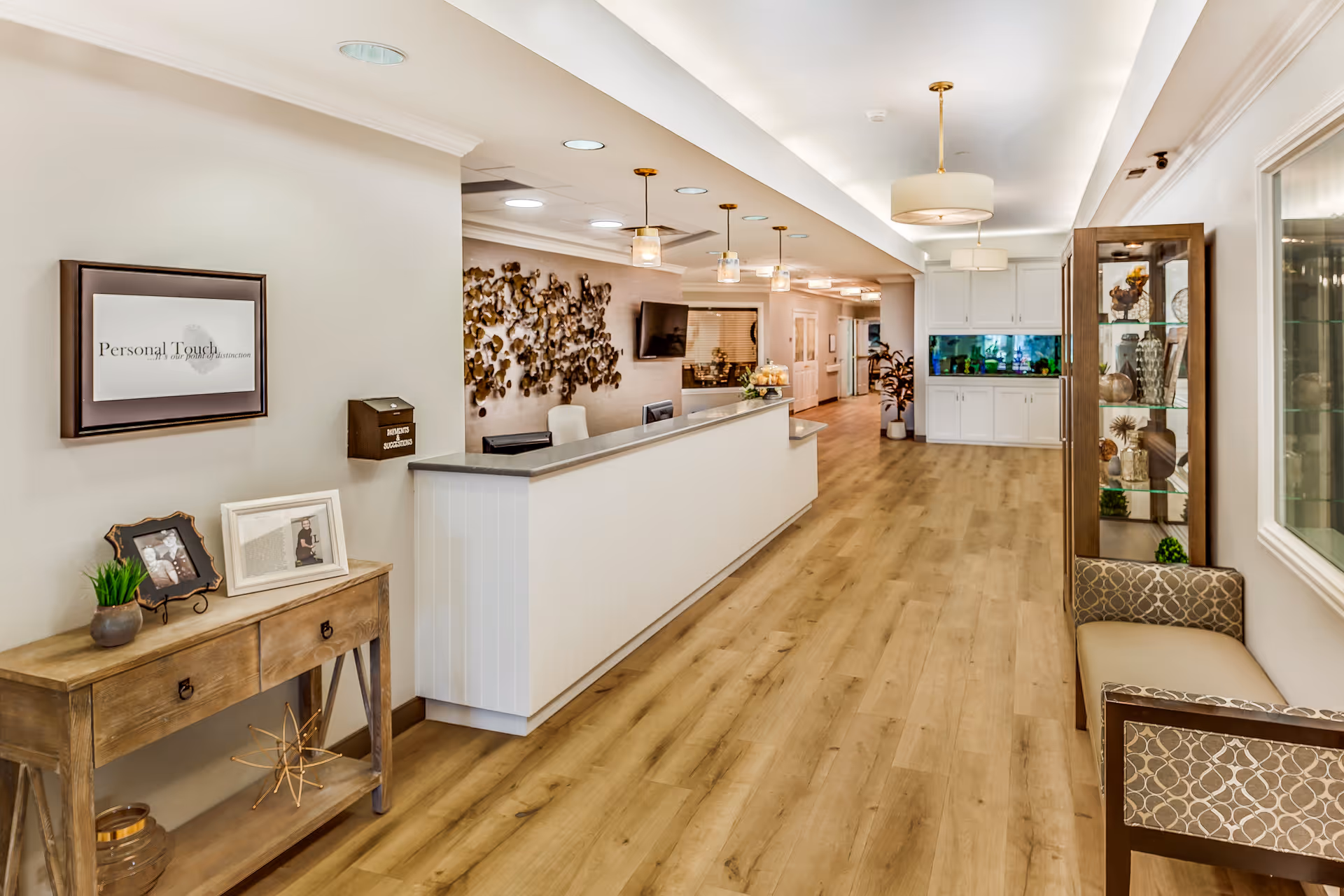A bright and spacious reception area with a long white front desk, wooden flooring, and modern pendant lights hanging from the ceiling. To the left, there is a wooden console table with framed photos and a small plant. On the right, there is a cushioned bench with patterned upholstery and a glass display cabinet. The far end of the room features white cabinetry and a large fish tank. The walls are decorated with a metal art piece and a wall-mounted TV.